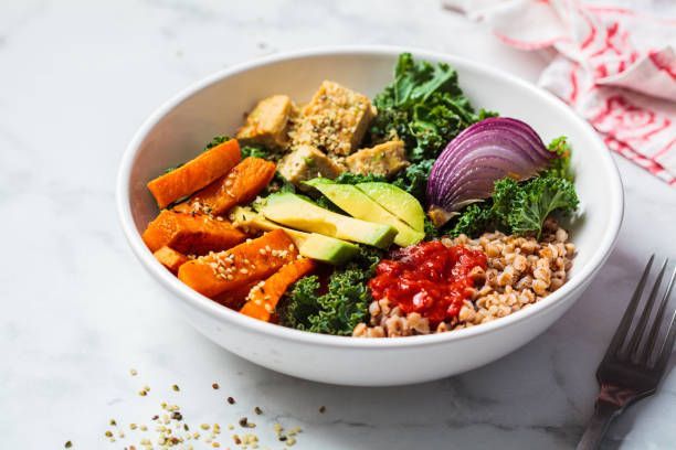 White bowl filled with a colorful vegan bowl: tofu, sweet potato, kale, avocado, red onion, and buckwheat groats.