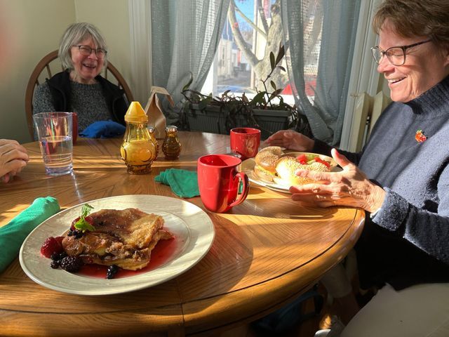 Two people at a sunny table, one holding food. Pancakes with berries and coffee cups.
