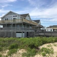 Large gray beach house with wraparound porch behind sandy dunes and grass under a cloudy sky