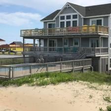 Gray beach house with large decks behind a sandy dune and wooden fence near the shore