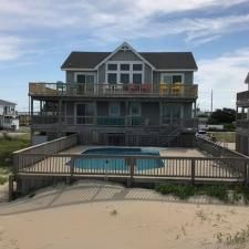 Beachfront house with a blue pool, wooden deck, and sand dunes under a cloudy sky