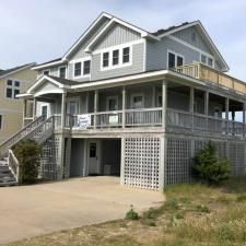 Gray beach house on stilts with wraparound porch, white trim, and a driveway under the elevated structure