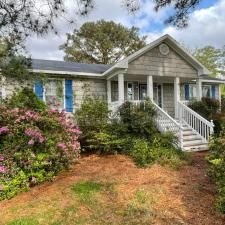 Single-story house with white siding, blue shutters, front porch, and steps surrounded by greenery and flowers
