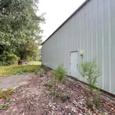 Long gray metal building beside a wooded path, with a small door and overgrown brush in front