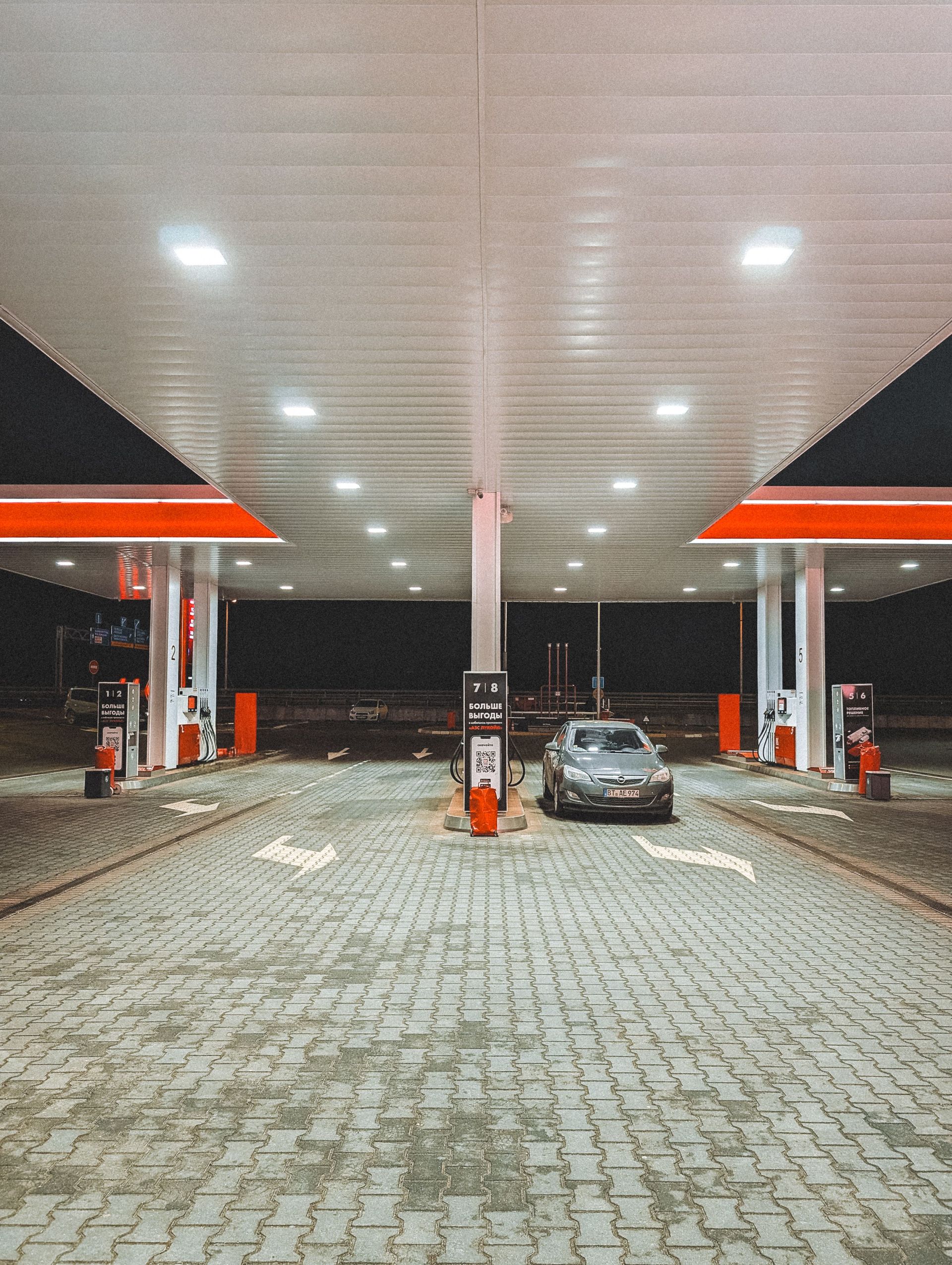 Nighttime gas station forecourt with a parked car under bright canopy lights and red-orange accents.