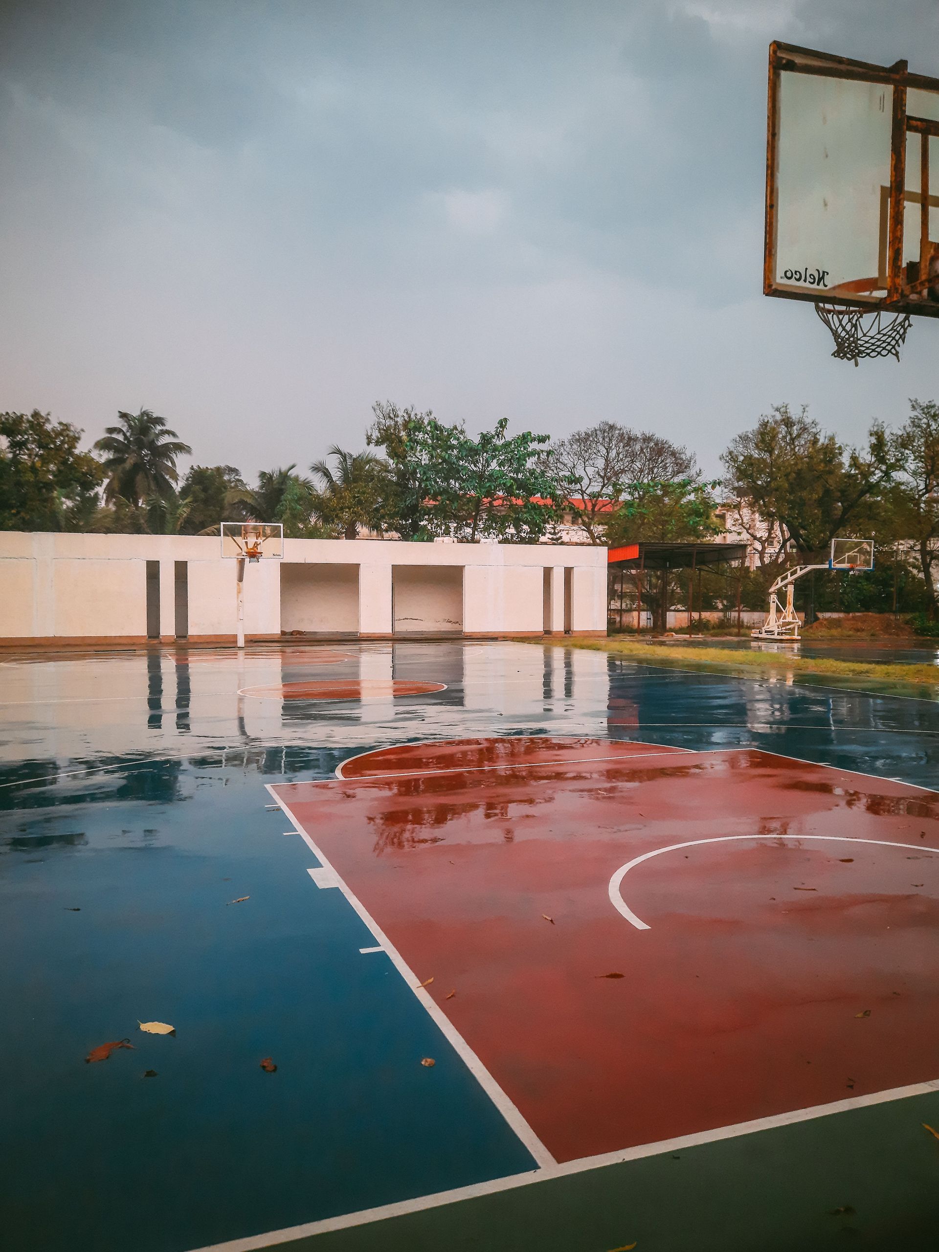 Empty outdoor basketball court with a wet red-and-blue surface and hoop under an overcast sky