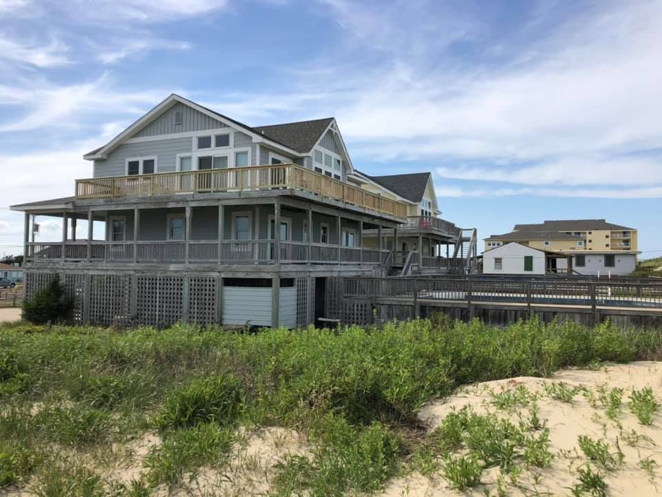 Raised beach house under construction with wraparound porch and gray siding in a sandy coastal lot