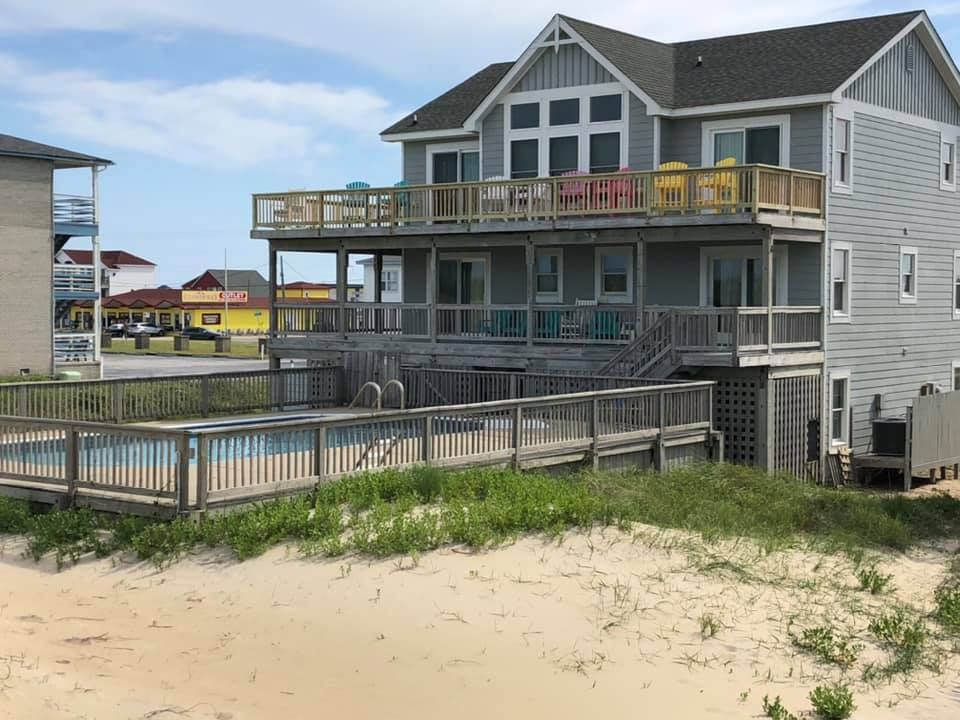 Beach house with gray siding, wraparound deck, and fenced pool area beside sandy dunes