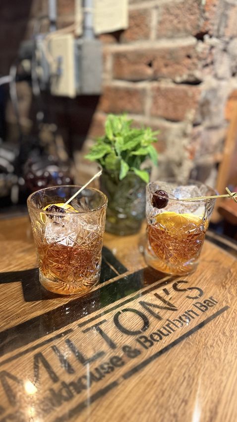 Two cocktails on a wooden bar, with a small plant and brick wall behind them.