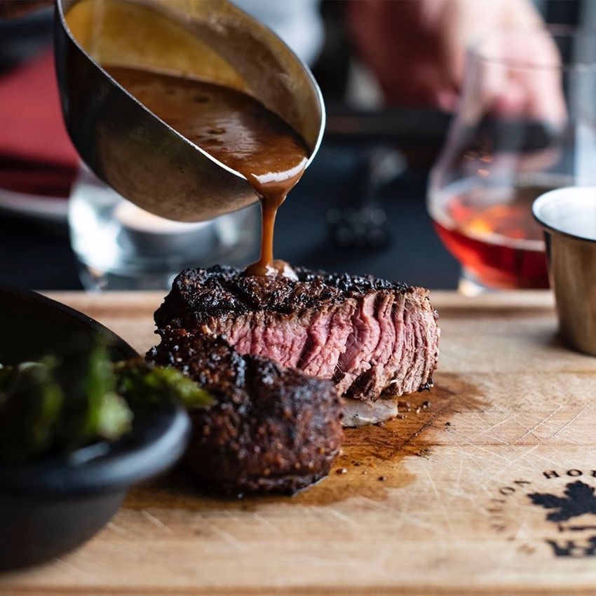 Steak being sliced on a wooden board, with sauce being poured over it in a restaurant setting