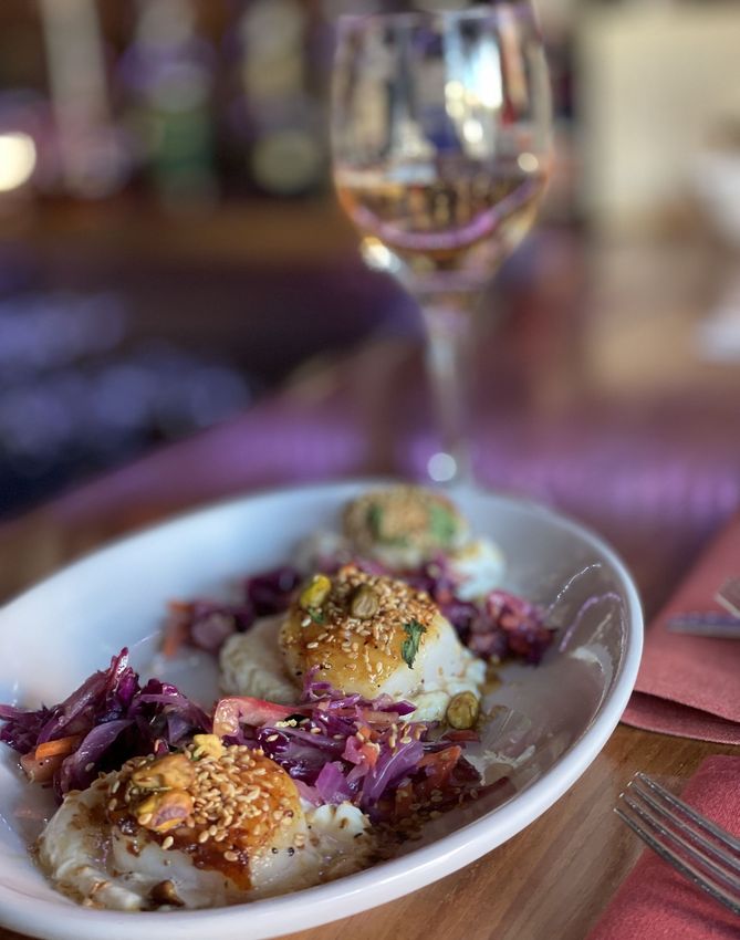 Plated scallop dish with red cabbage and mashed potatoes on a restaurant table, with a wine glass in the background