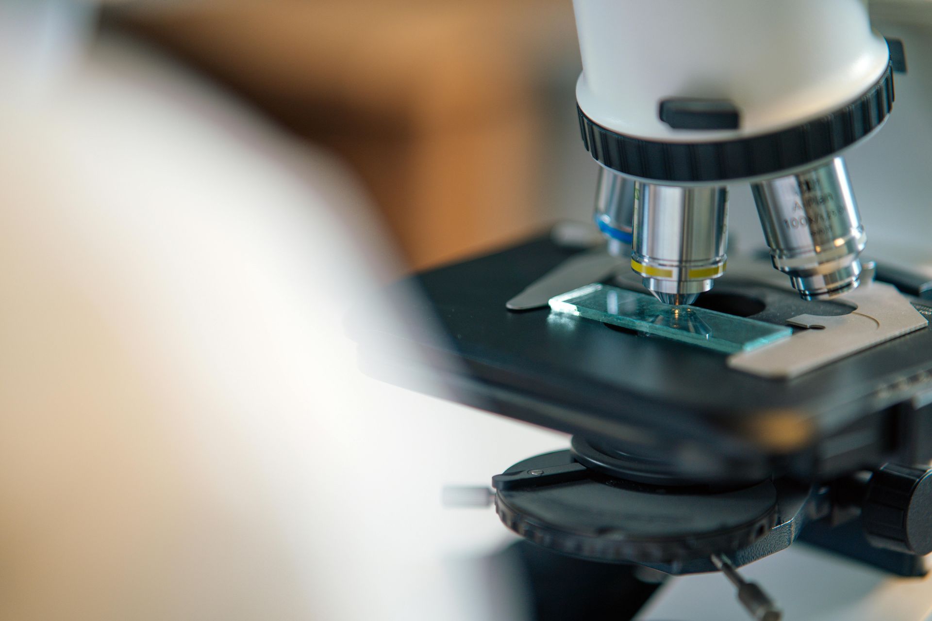 Donna in camice e occhiali protettivi che osserva un microscopio in un laboratorio.