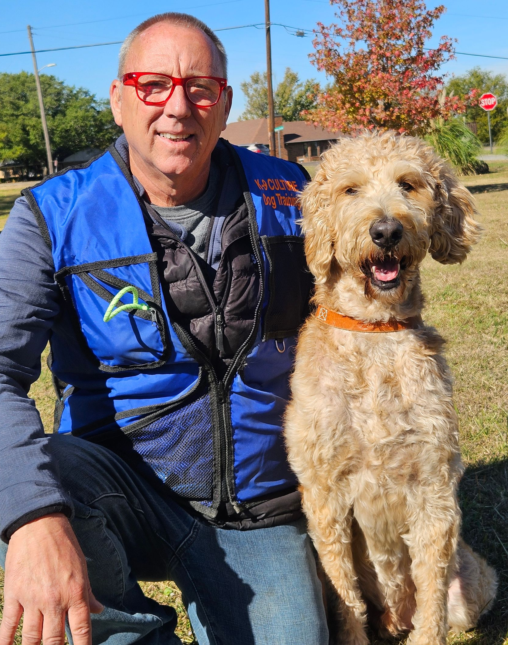 Man in blue vest kneels next to a golden doodle dog. Both smile outside on a sunny day.