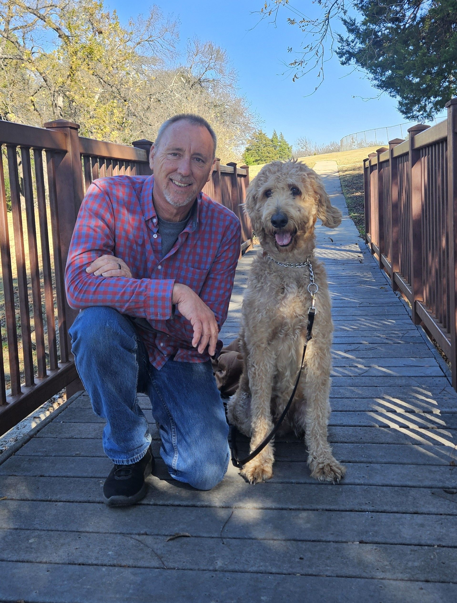 Man kneeling next to a golden doodle dog on a wooden bridge, both smiling. Sunny outdoors.