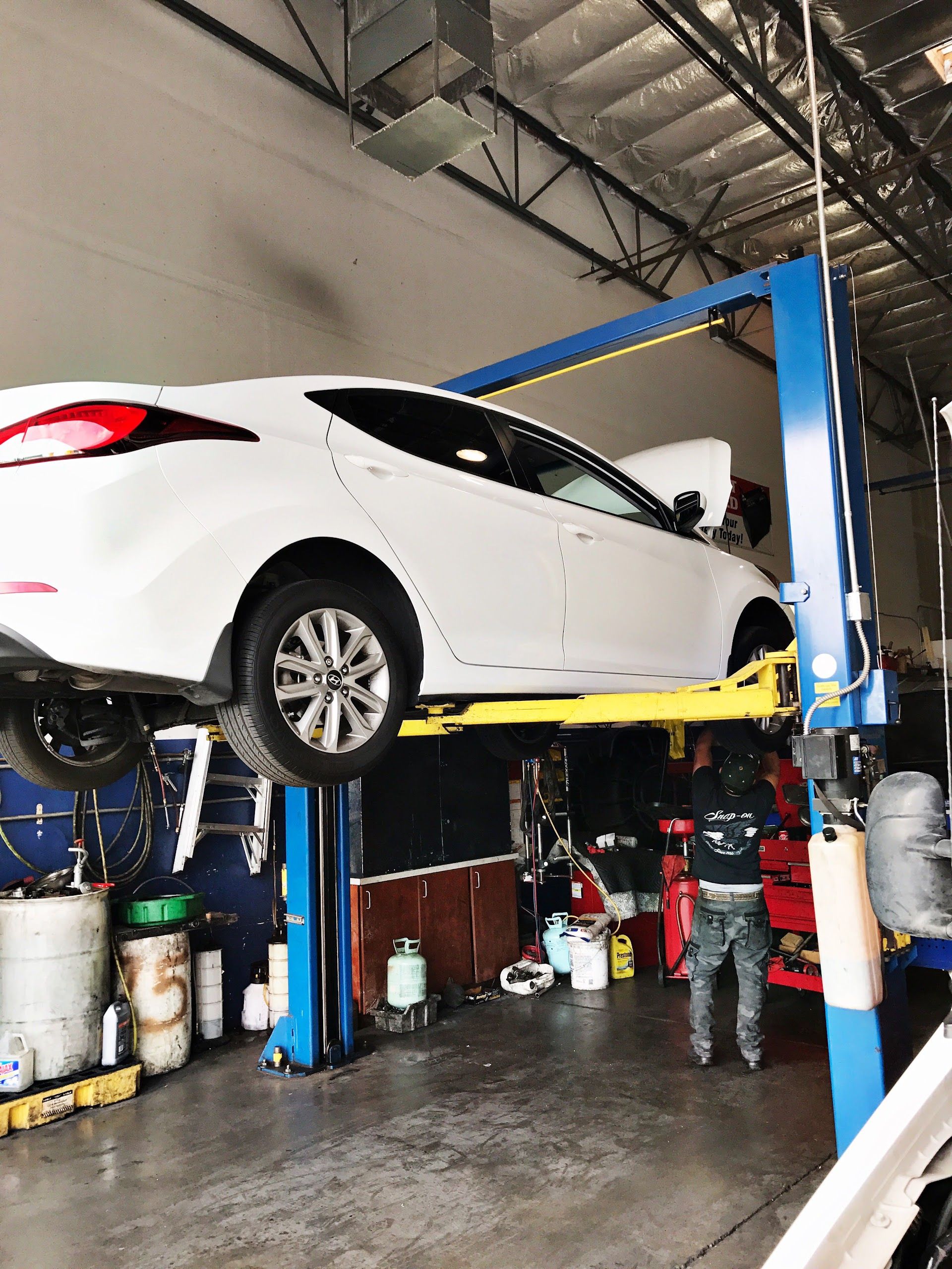 White car raised on a blue hydraulic lift in an auto repair shop with tools and supplies below | Velocity Motors