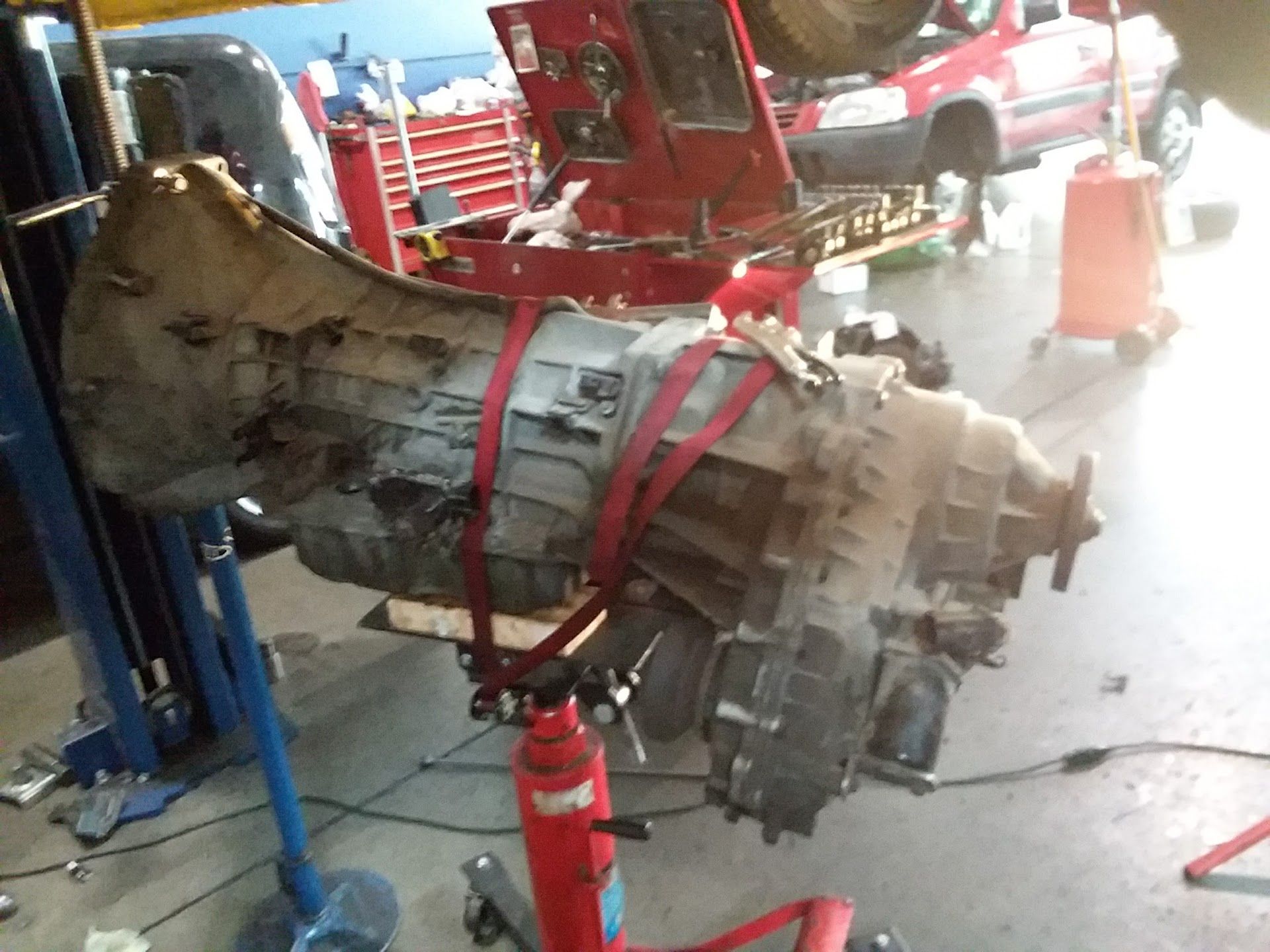 Damaged transmission suspended by straps in a workshop, with red equipment and tools in the background | Velocity Motors