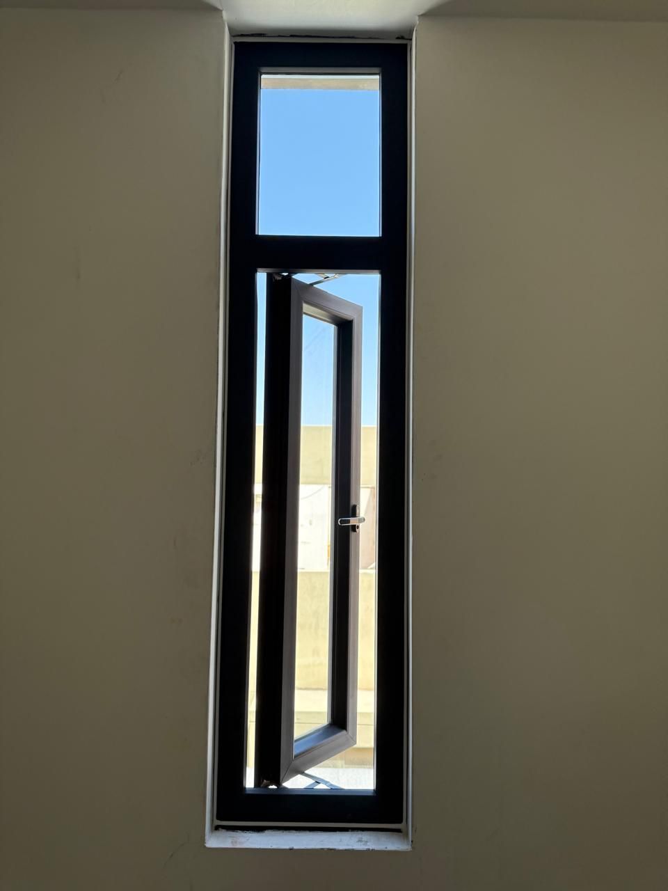 Un marco de ventana rectangular blanco con dos paneles, uno de los cuales está inclinado para dejar ver una pared beige y tejas en el tejado.