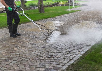 A person wearing protective gear uses a pressure washer to clean a stone-paved walkway next to a grassy lawn.