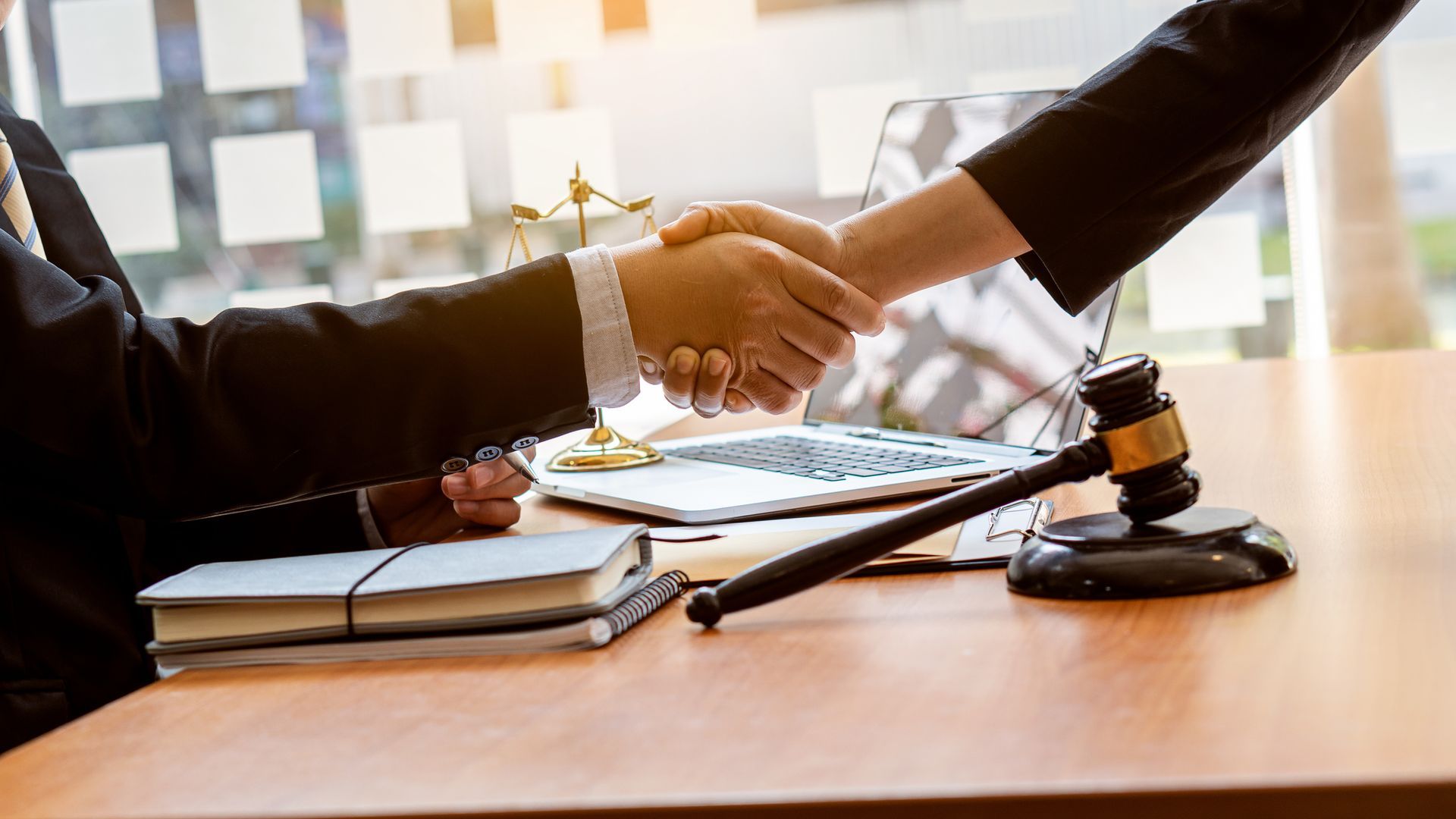 Two people shaking hands over a table with a gavel, legal books, and a laptop. Two people shaking hands over a table with a gavel, legal books, and a laptop.