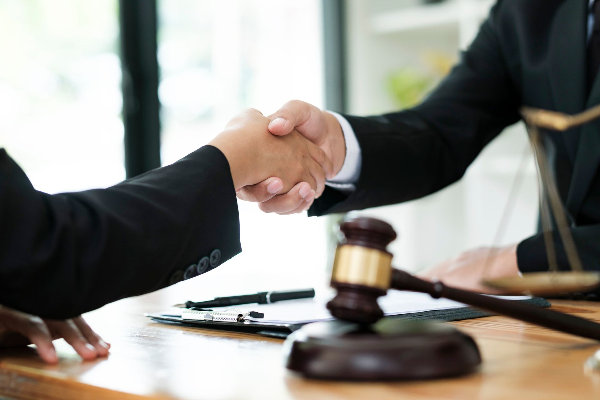 Two people in suits shaking hands at a table, gavel and scales of justice in foreground.