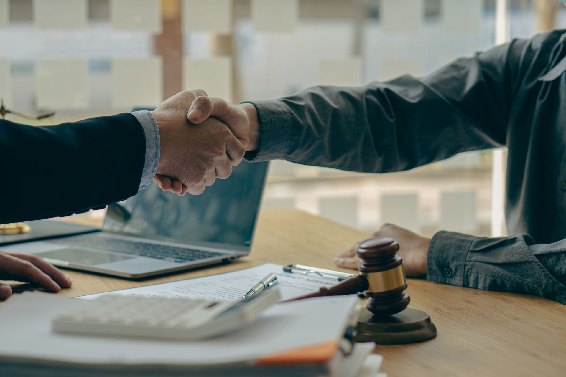 Two people shaking hands over a desk with documents, a gavel, and a laptop. Two people shaking hands over a desk with documents, a gavel, and a laptop.