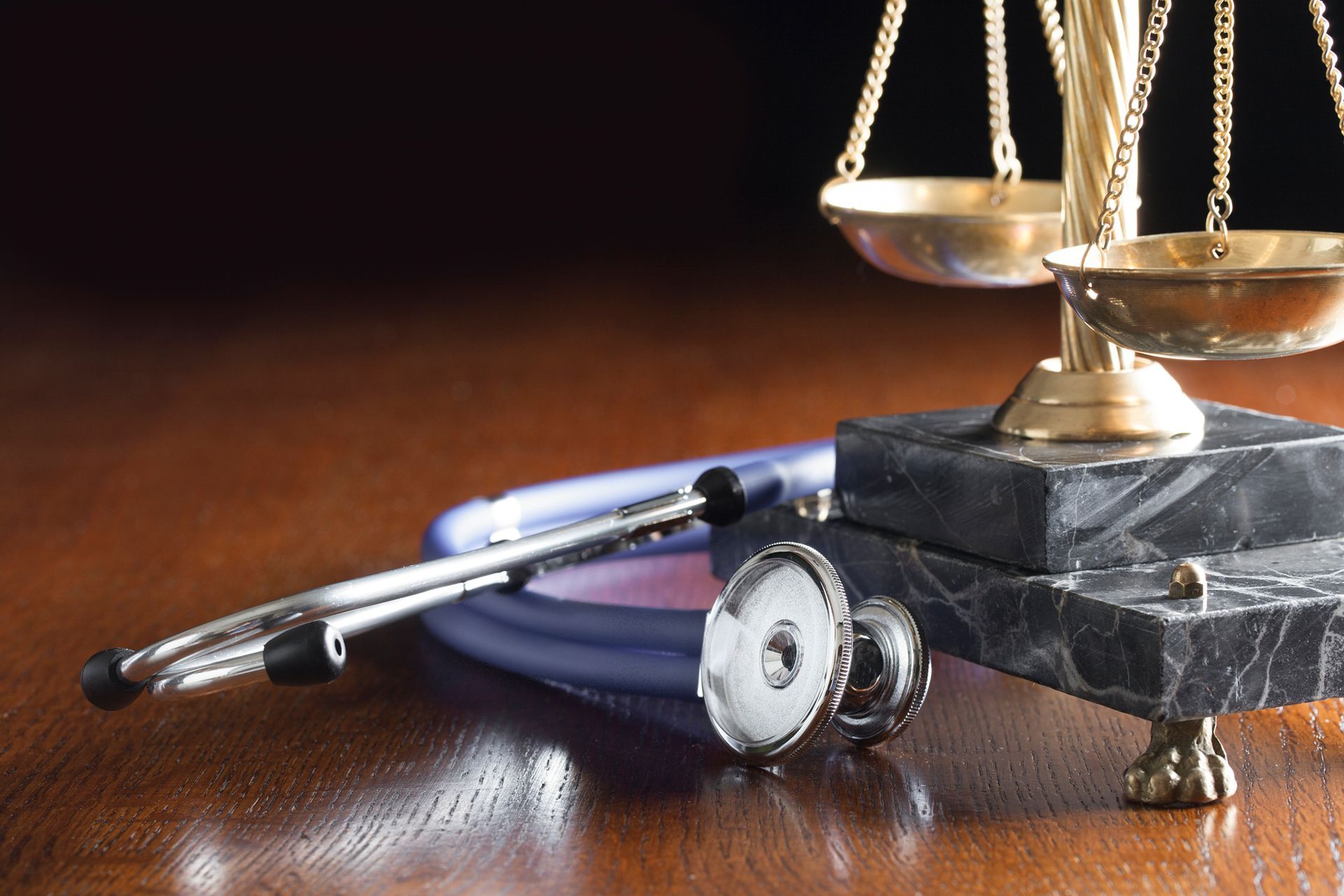 A stethoscope rests near a justice scale on a wooden surface, suggesting medical law.