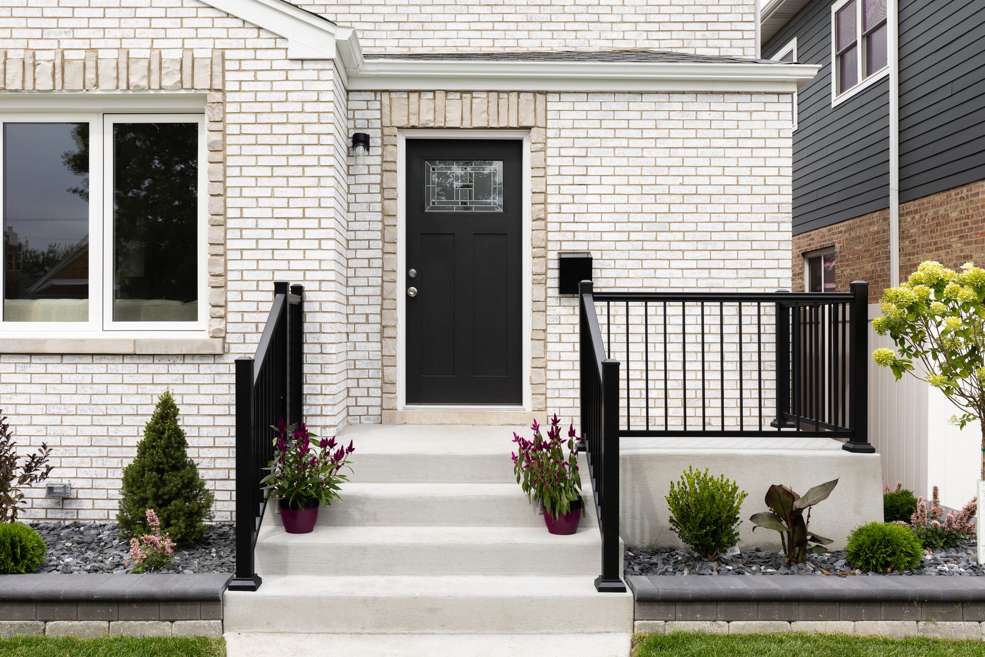 Front entrance of a white brick house with black railings, steps, and potted flowers.