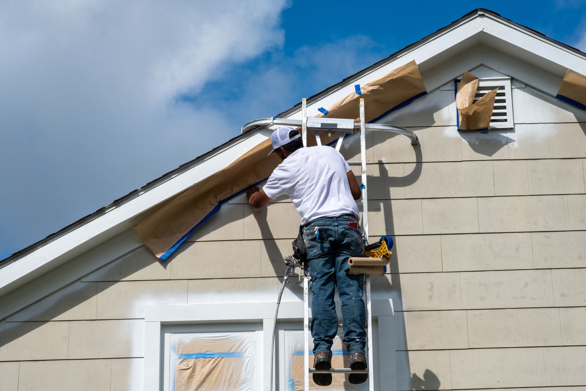 Worker painting house trim from a ladder under a blue sky