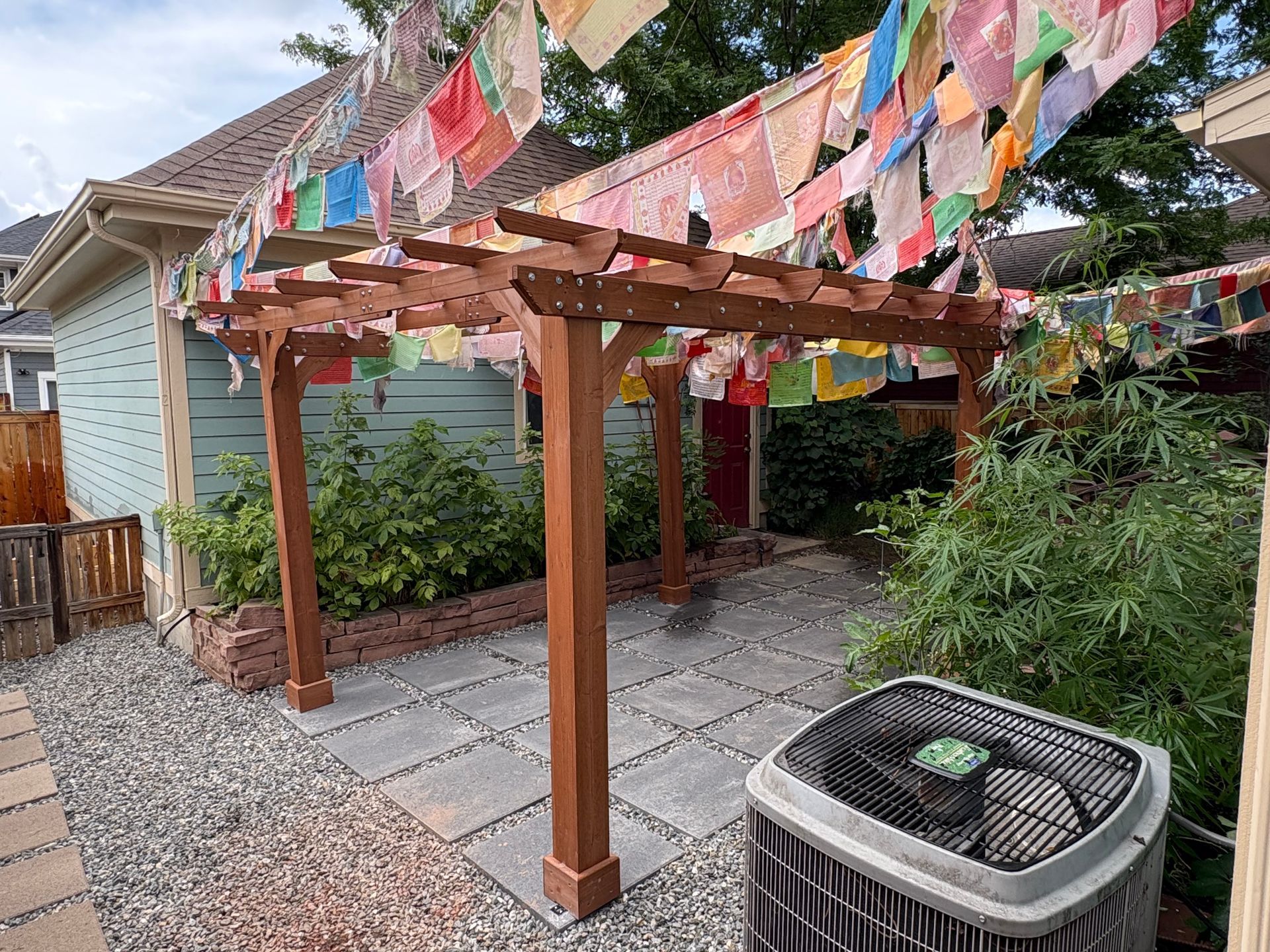 Wooden pergola in a backyard, strung with colorful prayer flags. A/C unit in the foreground.