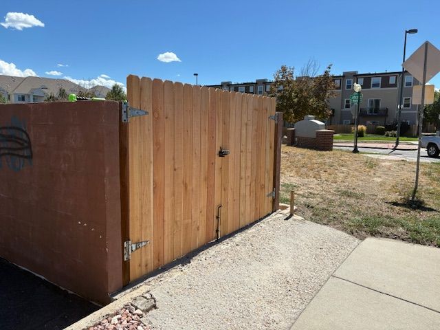 Wooden gate in a tan fence, adjacent to a brown enclosure. Sunny day with buildings in the background.