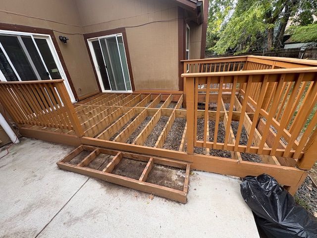 Deck renovation in progress; wooden deck frame exposed, next to patio and house with sliding glass door.