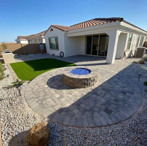 Patio with a fire pit, artificial grass, and a white house under a blue sky.