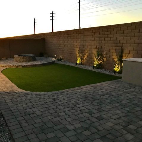 Backyard with brick wall, green grass, patio, fire pit, and plants lit by spotlights at sunset.