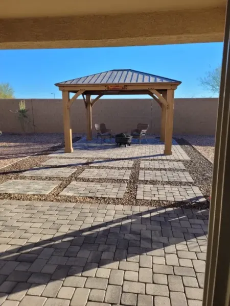 Backyard patio with gazebo, stone pavers, and a dark dog. Sunny day.