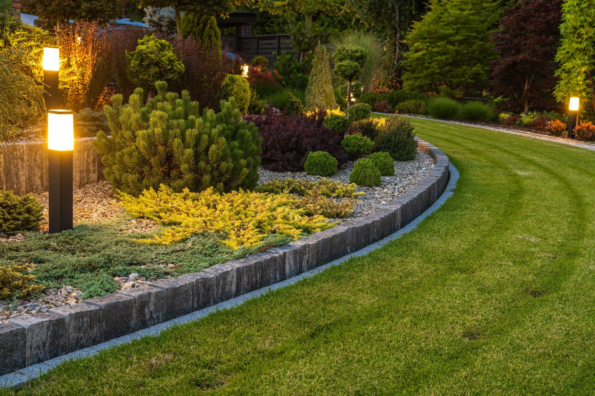 A garden bed with various plants is lit by outdoor lamps along a curving lawn path.