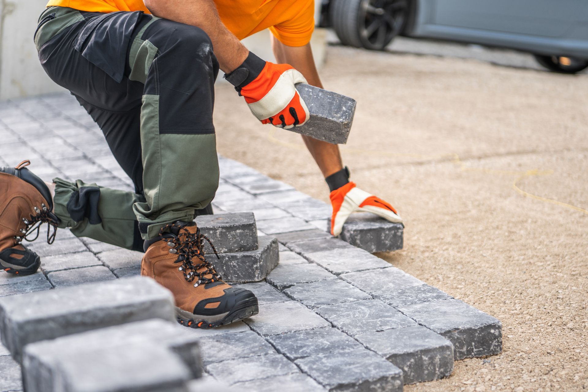 Construction worker kneeling, laying gray paving stones on a driveway, wearing work gloves and boots.