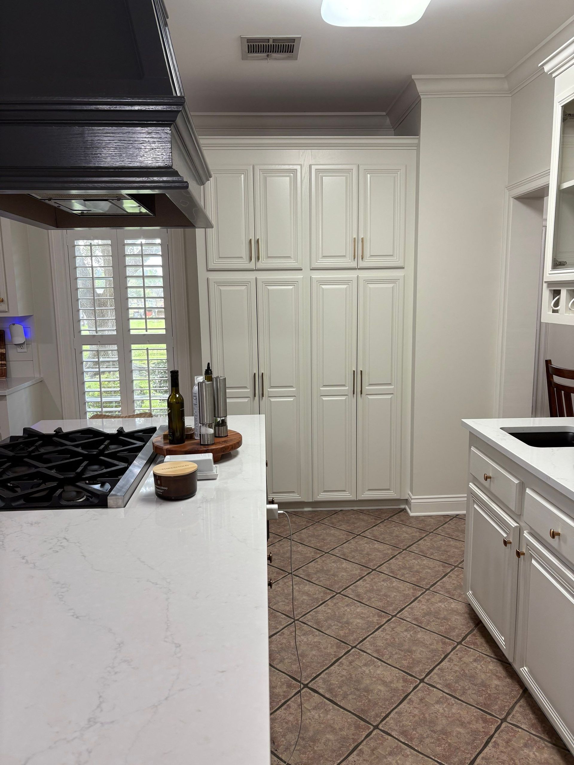 Kitchen with white cabinets, countertop, stove, and tall pantry. Tile floor. Window in the background.