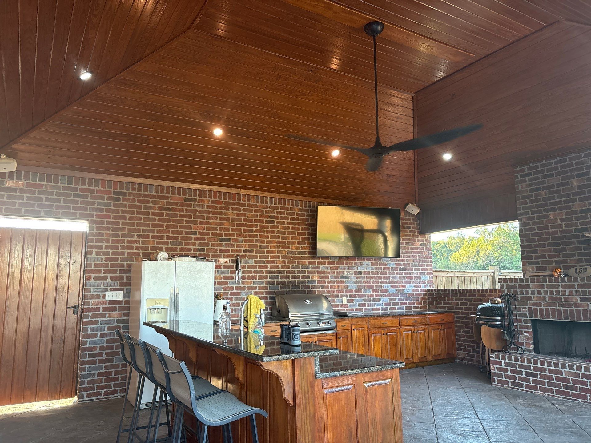 Outdoor kitchen with brick walls, wooden cabinets, and ceiling. Features a bar, grill, TV, and ceiling fan.