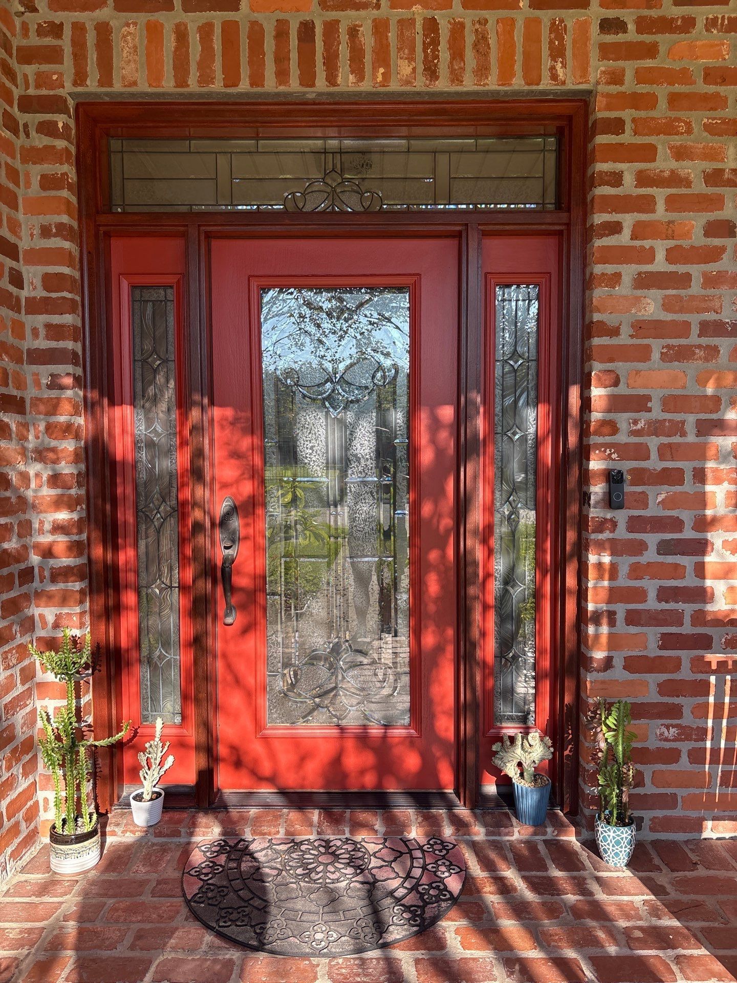 Red front door with glass panels on a brick house; plants and a doormat are present.