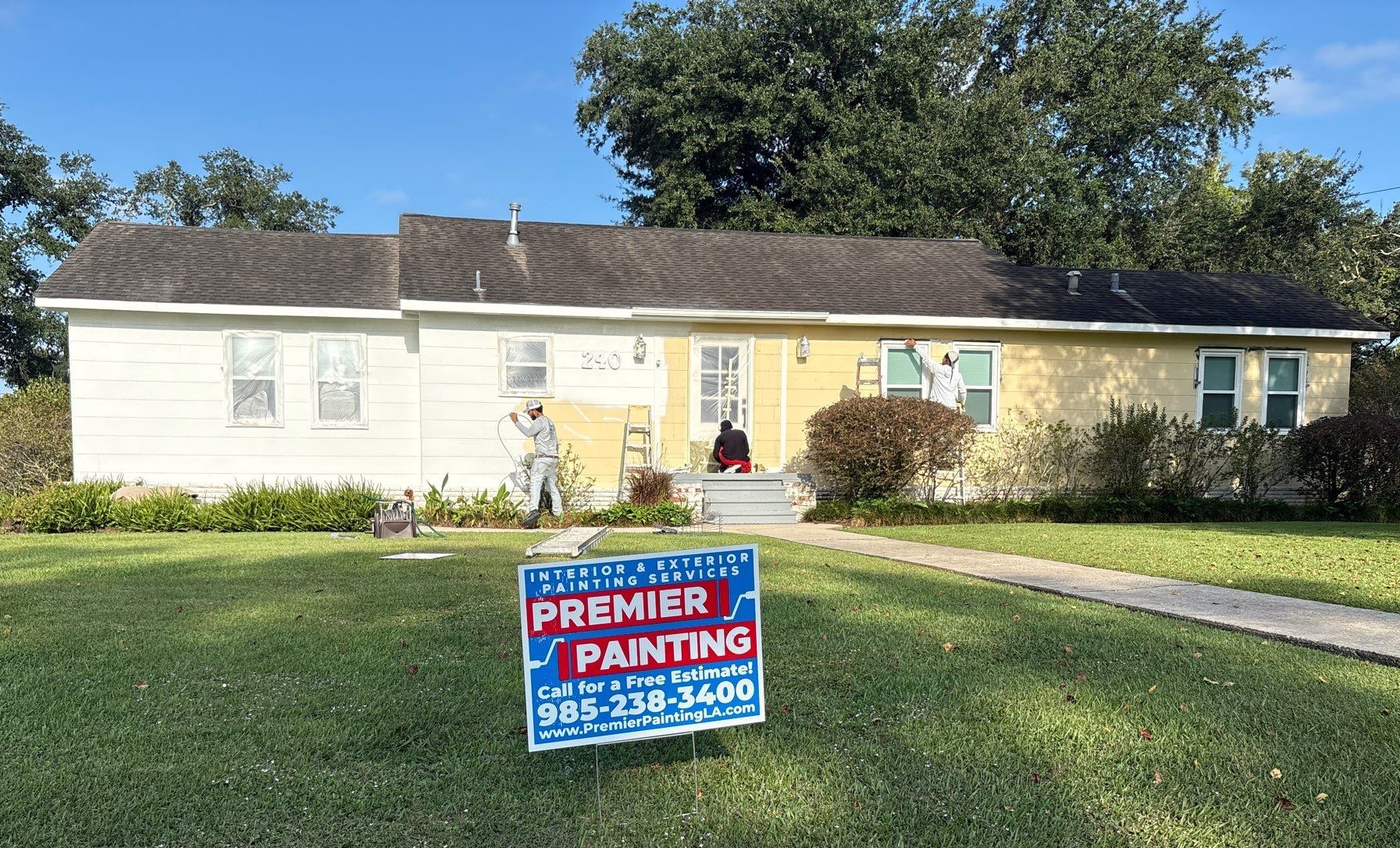 House being painted; one side white, the other yellow. Premier Painting sign on lawn, workers visible.