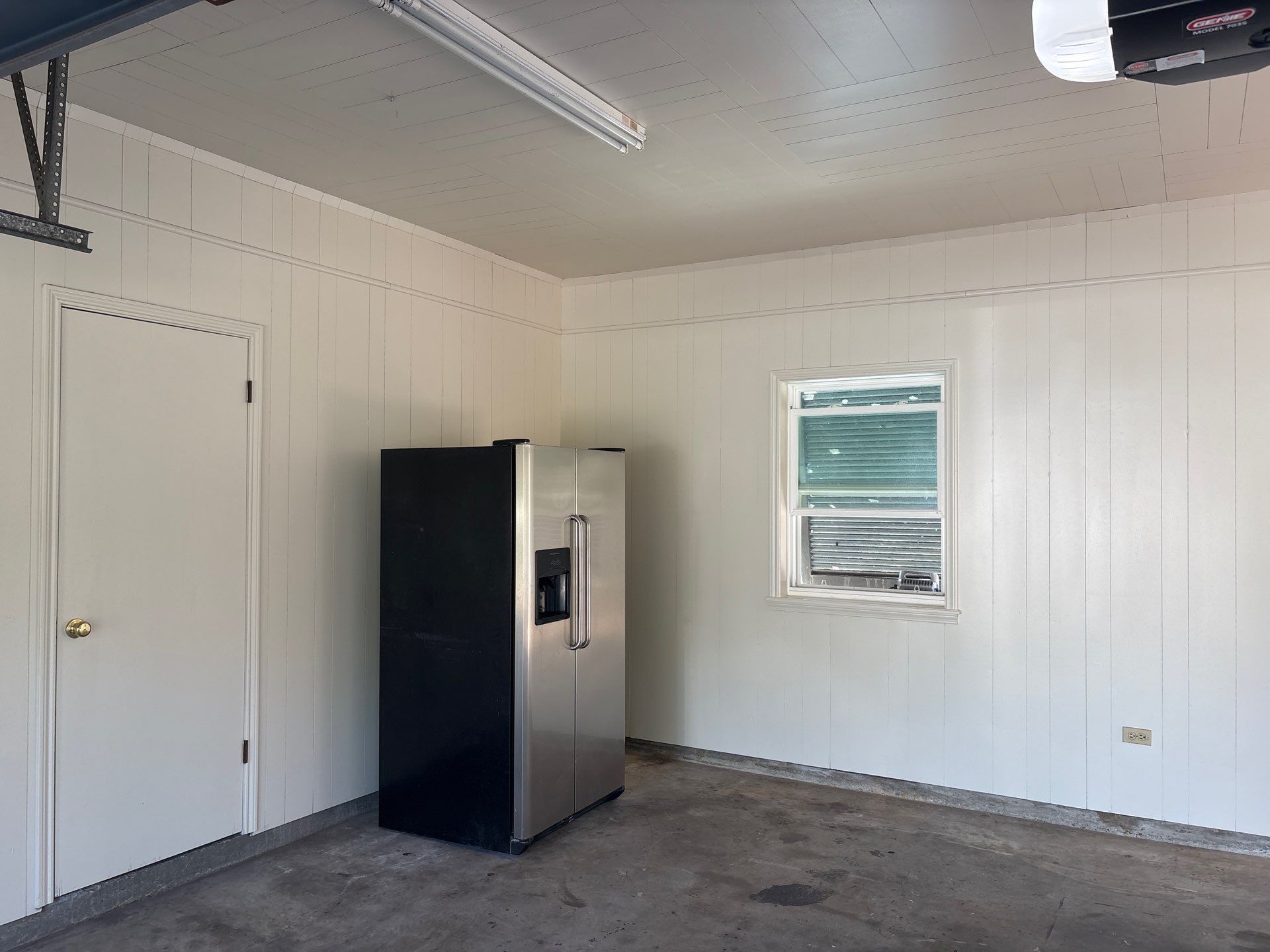 Garage interior with a refrigerator, door, and window. Walls are white, floor is concrete.