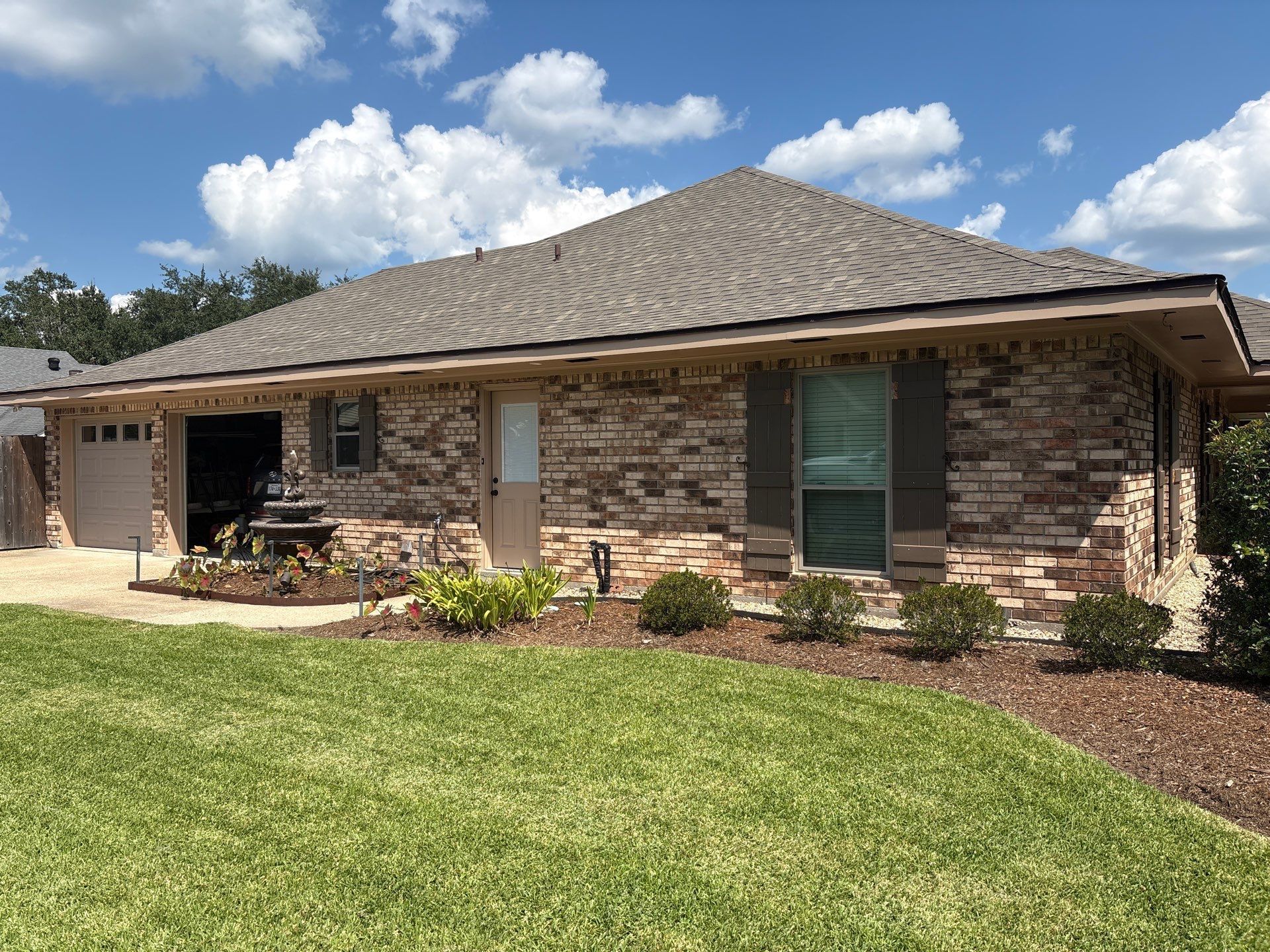 Brick house with brown roof, green grass, and small garden bed under a bright blue sky.