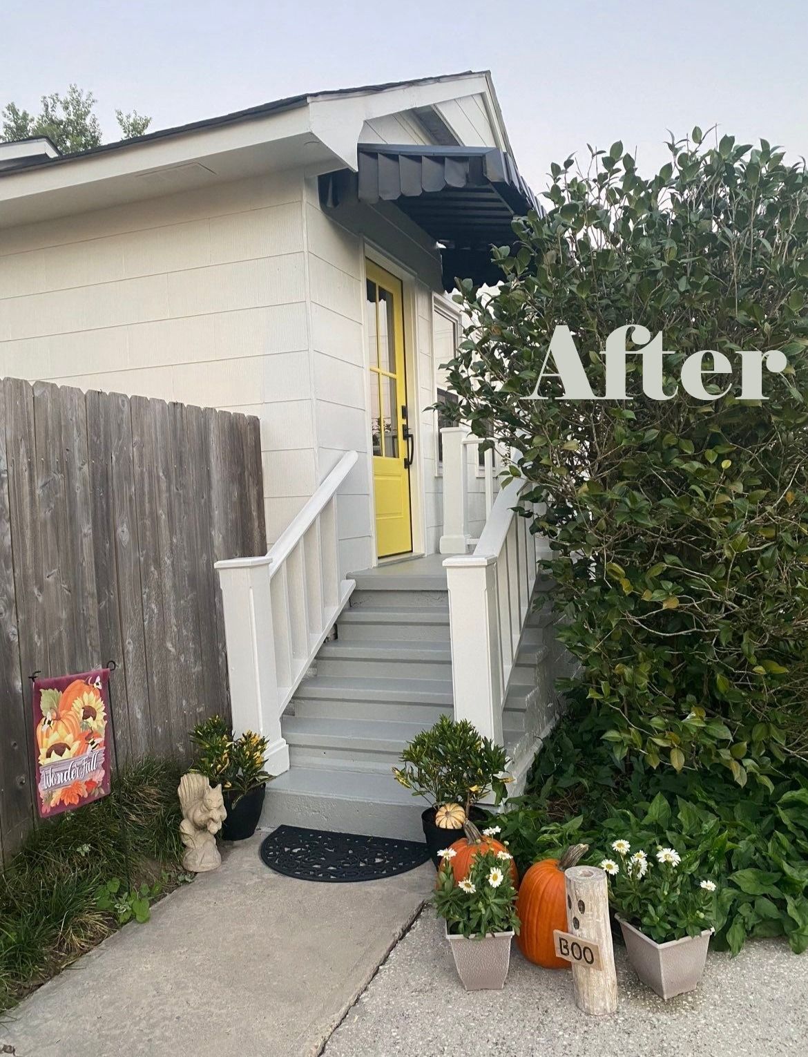 Curb appeal, updated entrance: White house with yellow door, gray steps, black awning, pumpkins, flowers, and wooden fence.