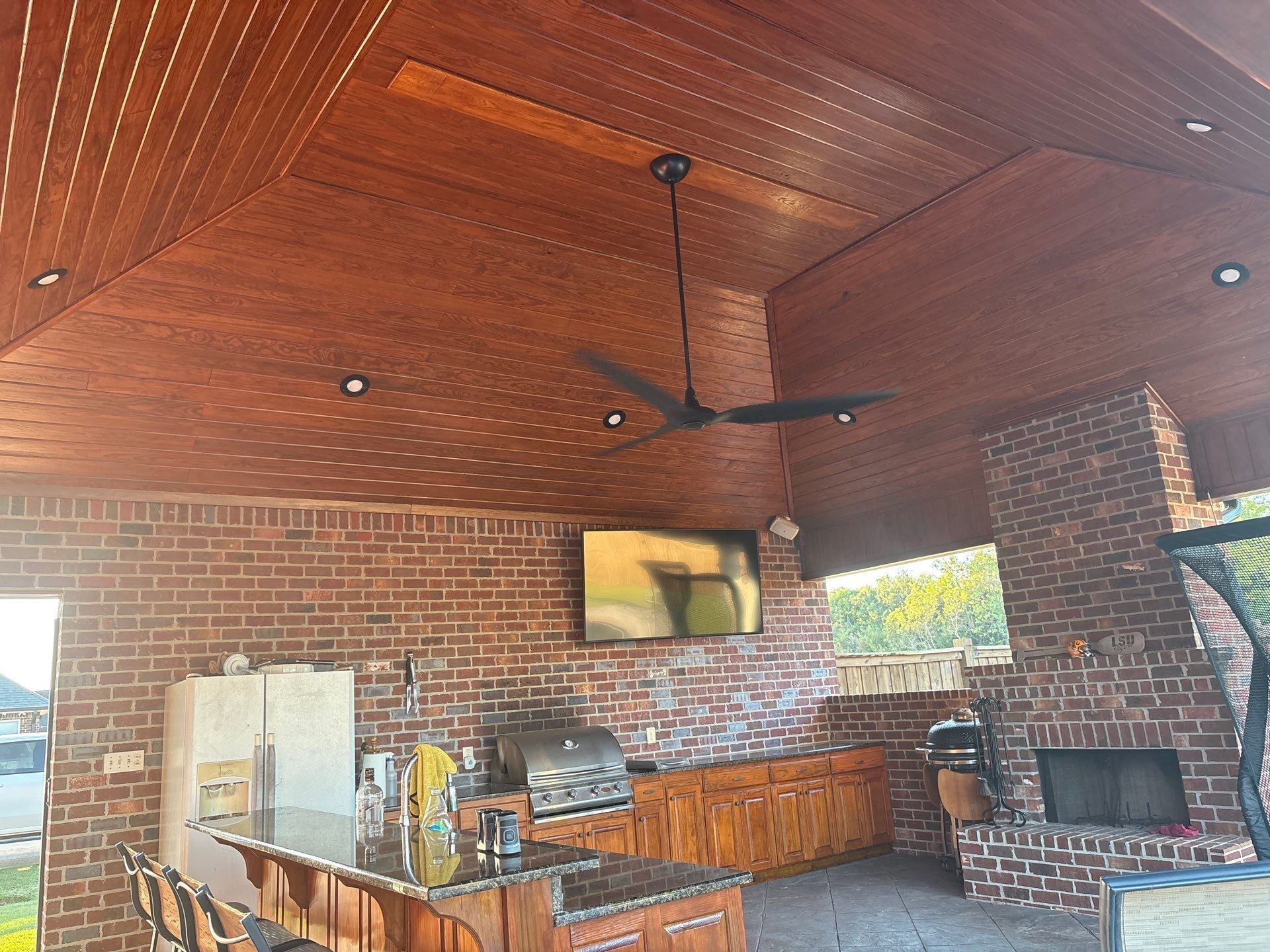 Outdoor kitchen with brick walls, wood ceiling, and appliances including a grill, refrigerator, and TV.
