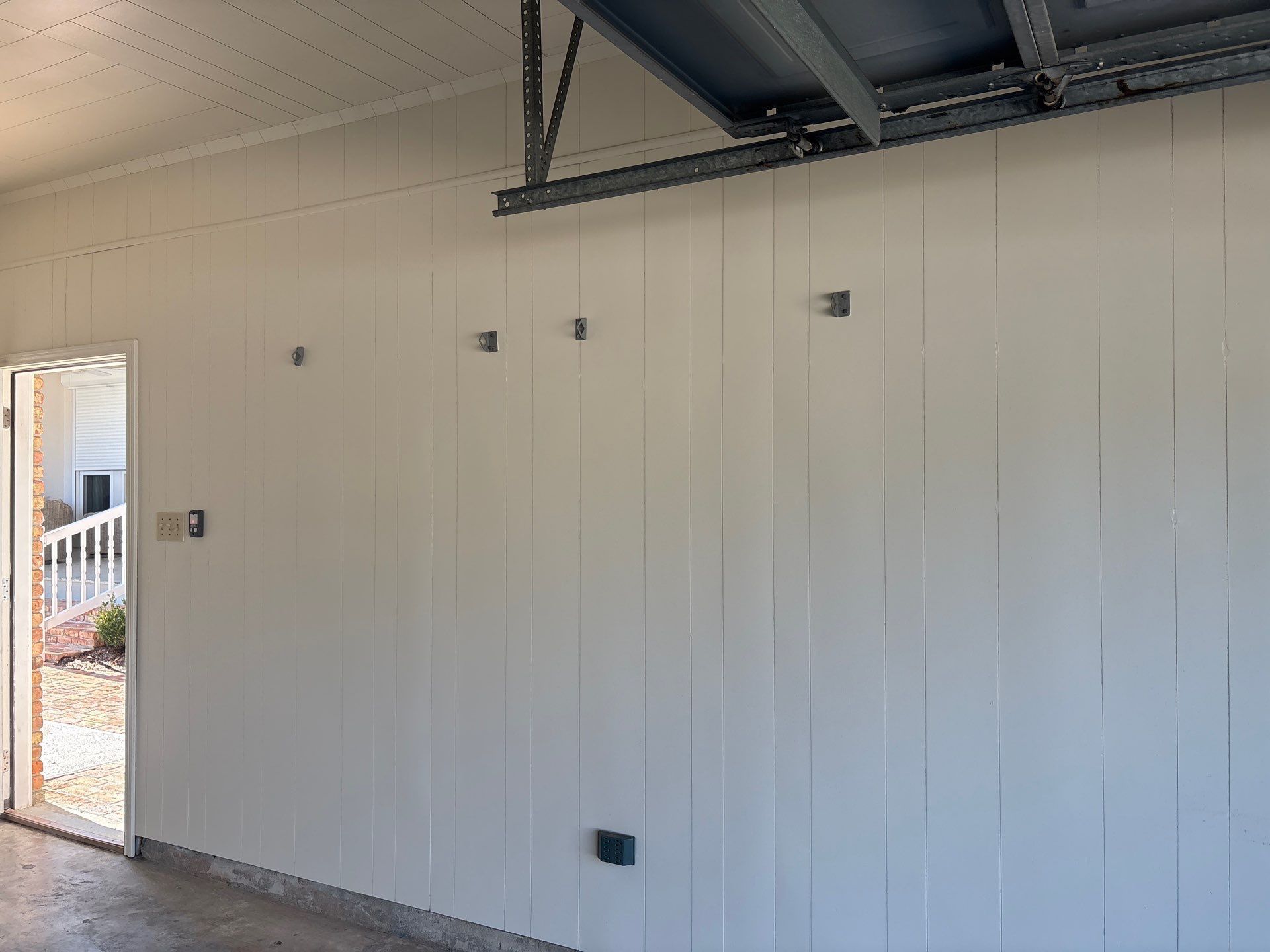White-paneled garage wall with four gray squares. Open door to outdoors on left. Metal garage door rail at top.