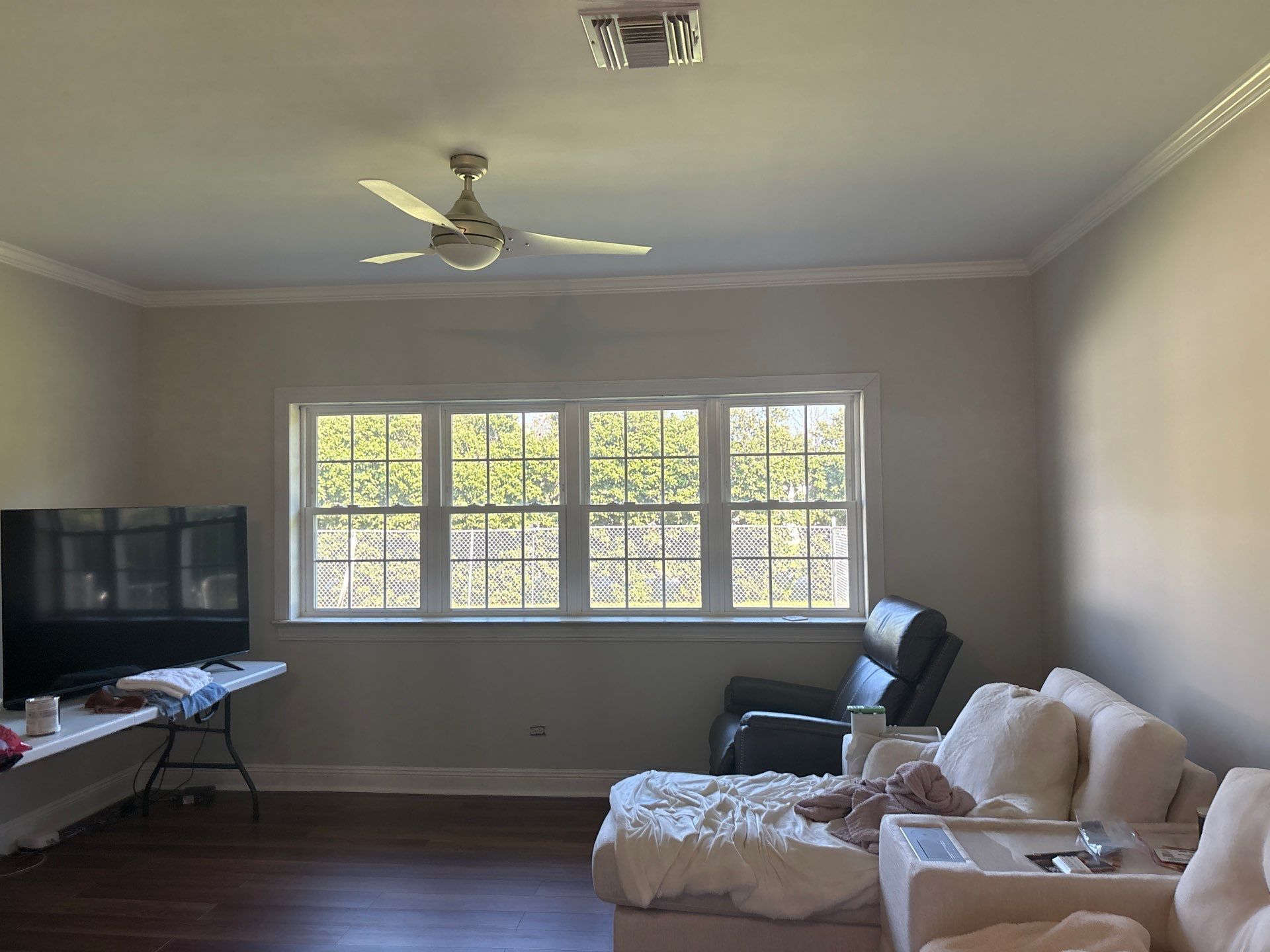 Living room with large windows, TV, sofa, and ceiling fan. Natural light fills the space; cream-colored walls.