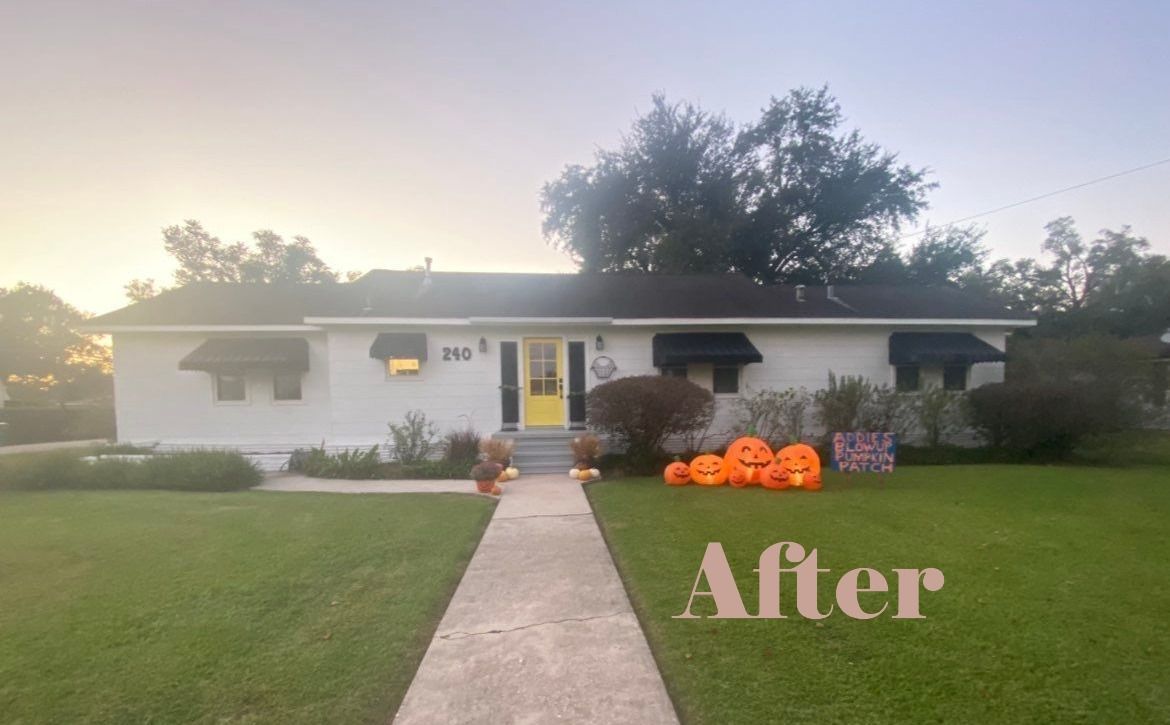 White house with yellow door, pumpkins, and a walkway on a grassy lawn.
