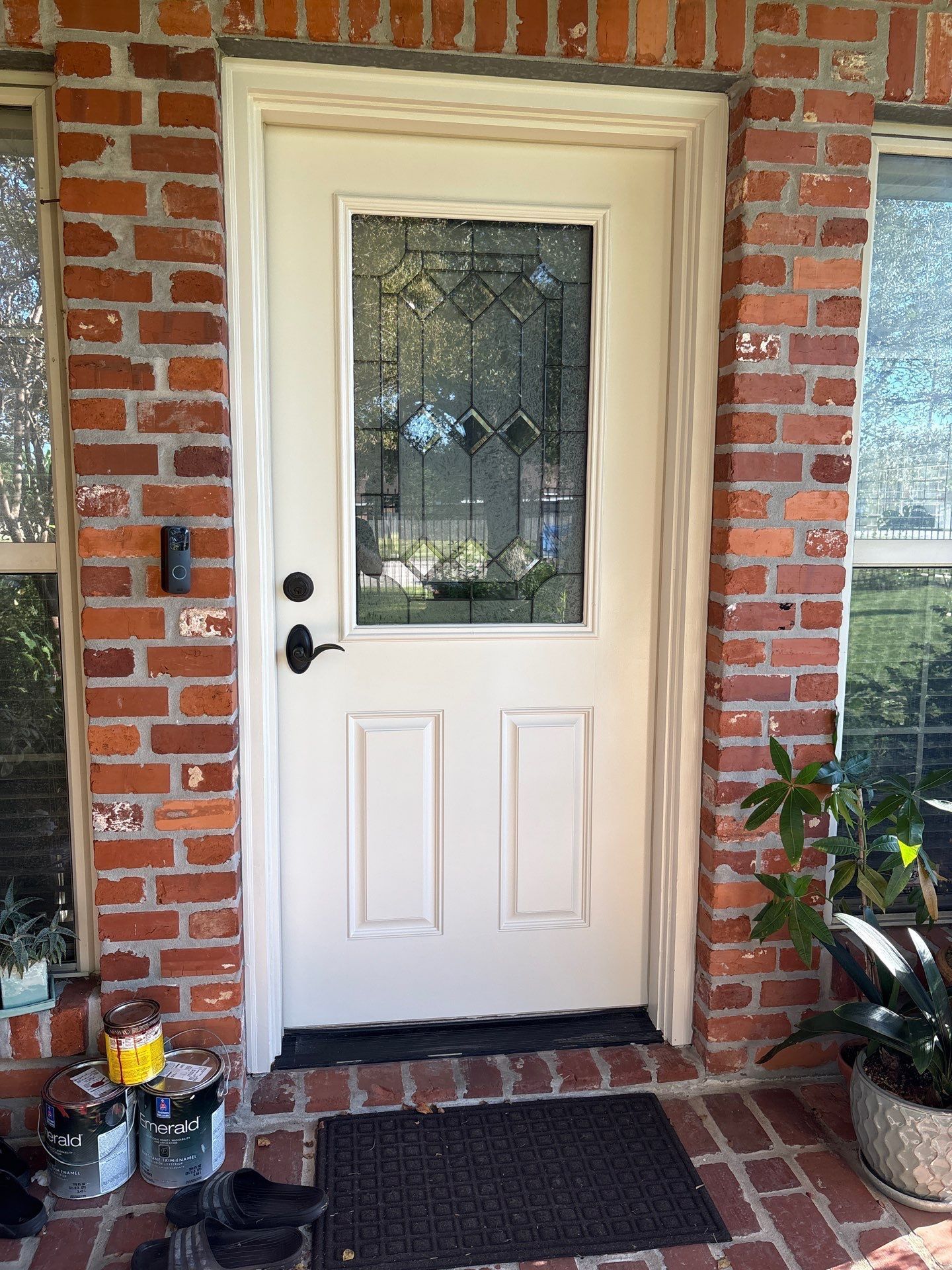 Cream-colored front door with decorative glass panel, flanked by brick walls and windows. Black doormat and potted plants.