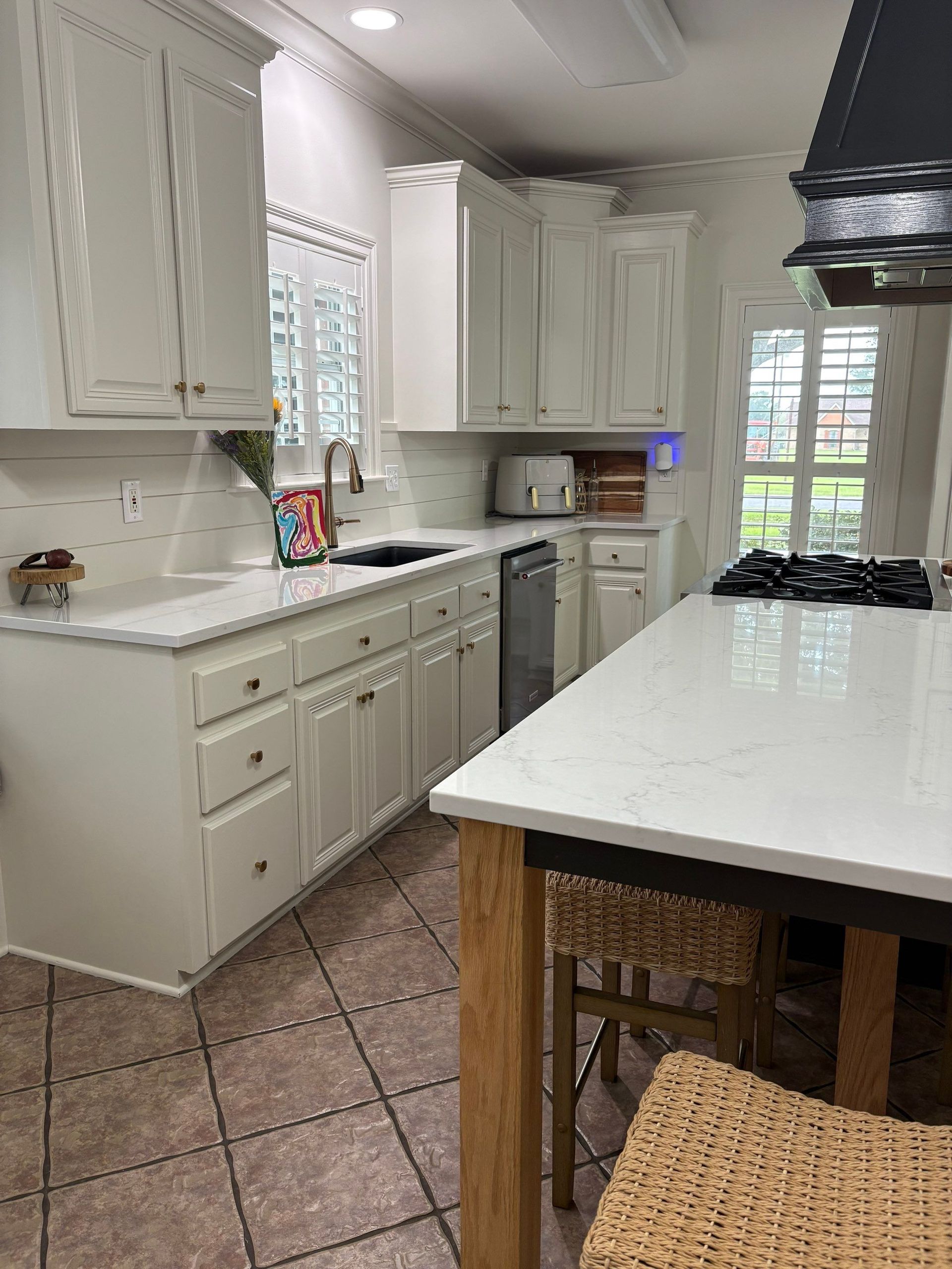 White kitchen with island, cabinets, and appliances. Brown tile floor, window, and wicker bar stool.