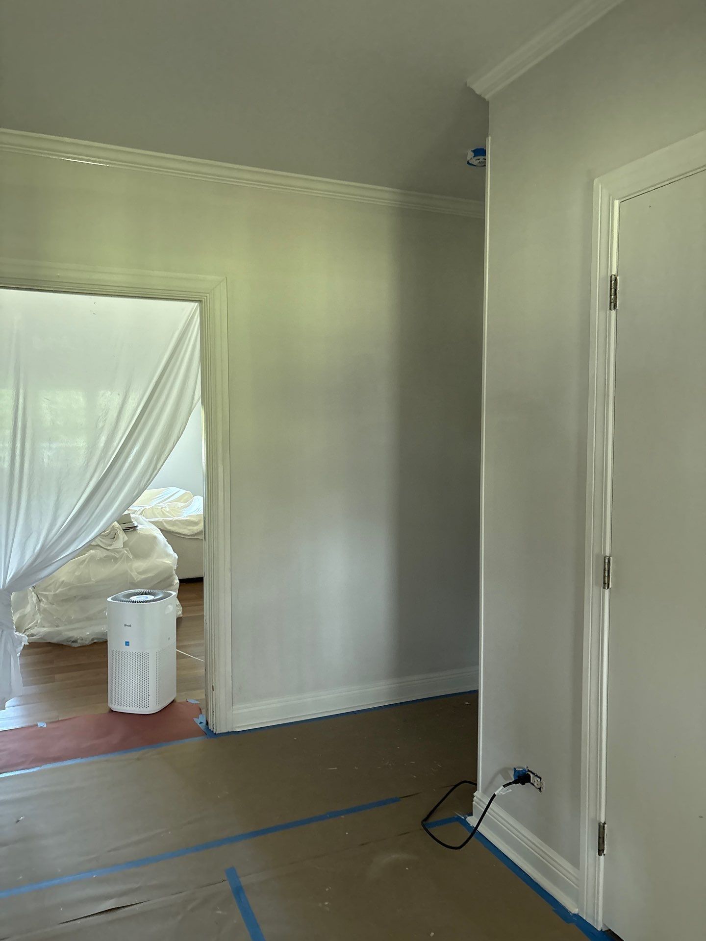 Room interior during painting. Light grey walls, white trim and doorway. Protective sheeting covers room.